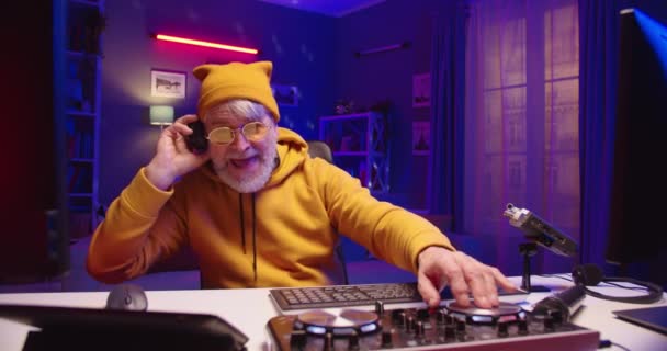 Close up portrait of joyful Caucasian old bearded male DJ listening to music in headphones working on console mixer mixing new music on vinyl plate sitting at table in room at home studio, neon lights — Stock Video
