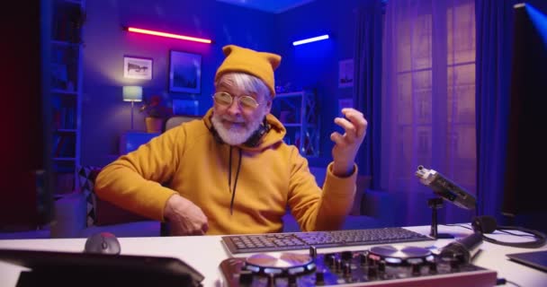 Portrait of Caucasian cheerful old grey-haired professional man DJ in yellow hat working on console mixer mixing music using vinyl plate having fun in good mood sitting in apartment with neon lights — Stock Video