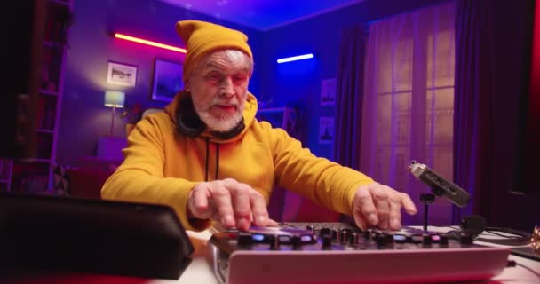 Glücklicher, stilvoller kaukasischer Senior-DJ, der im heimischen Aufnahmestudio Musik spielt. Großaufnahme eines alten Mannes, der Geräusche auf einem Mischpult mixt, der in einem Zimmer in einer Wohnung in Quarantäne sitzt. Hobby-Konzept — Stockvideo