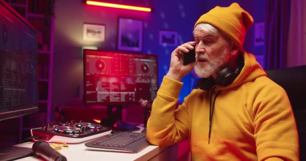 Közelkép kaukázusi elfoglalt idős férfi dj sárga kalapban ül a sötét szobában beszél okostelefon hívás és keverés hangulat zene vinil lemez mixer konzol dolgozik a számítógép, dj koncepció — Stock videók