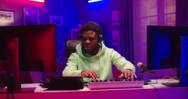 Portret van een knappe jongeman DJ die thuis opnamestudio speelt in neonlicht. Studenten leren creatieve sfeermuziek schrijven. Afro-Amerikaanse man componeren geluiden op mixer console — Stockvideo