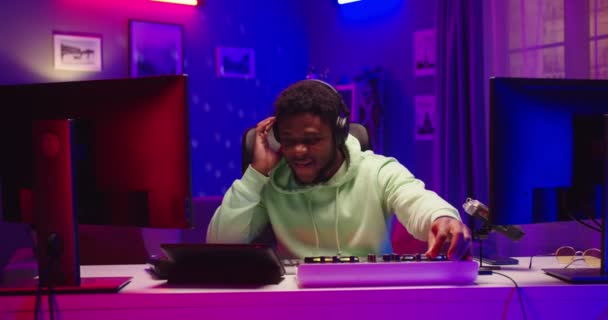 Portrait of funny joyful young African American male DJ playing music on mixer console and vinyl plate sitting in room at home recording studio in neon light smiling in good mood, profession concept — Stock Video