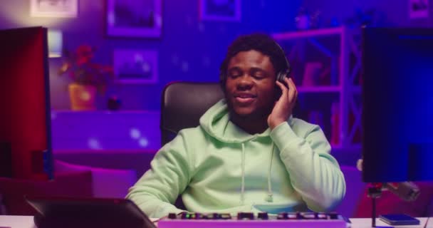 Close up portrait of happy African American young handsome man DJ composing music on mixer console and listening to song in headphones sitting in room in neon light streaming live, musician concept — Stock Video