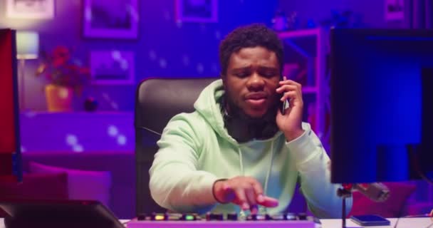 Молодой афроамериканец счастливый красавчик-ди-джей, создающий музыку, сочиняющую ритмы на микшерной консоли, разговаривая на смартфоне, сидя в домашней студии в неоновом свете — стоковое видео