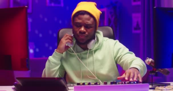 Κοντινό πορτραίτο του νεαρού όμορφου Αφροαμερικανού DJ που παίζει μουσική στην κονσόλα του μίξερ με τη χρήση πίνακα μίξης και πινακίδας βινυλίου ακούγοντας ήχο στα ακουστικά που κάθονται στο δωμάτιο με νέον φως — Αρχείο Βίντεο