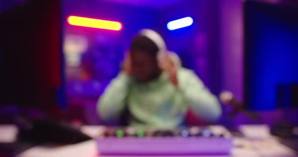 Camera approaching emotional happy African American young male dj playing music on mixer console sitting in neon lights at home recording studio. Close up of dj scratching vinyl creating beats — Stock Video