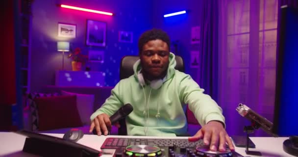 アフリカ系アメリカ人の若者の肖像DJがターンテーブルコンソールでクラブ音楽を演奏ネオンライトとマイクで部屋の暗いホームスタジオに座っているDJストリーミング音楽ライブとオンラインで話している — ストック動画