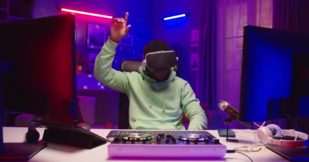 Joyeux jeune afro-américain positif DJ masculin assis dans la pièce sombre dans des néons portant des lunettes VR en utilisant la technologie futuriste. Homme en réalité virtuelle casque mélangeant la musique dans le studio d'enregistrement à domicile — Video