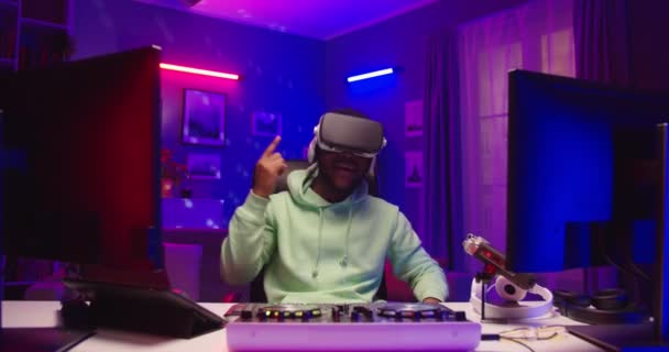 Fényképezőgép közeledik a fiatal afro-amerikai boldog srác DJ élvezi szórakoztató futurisztikus technológia visel VR headset ül neon fény és tánc ritmikusan. Férfi virtuális valóság szemüveg — Stock videók