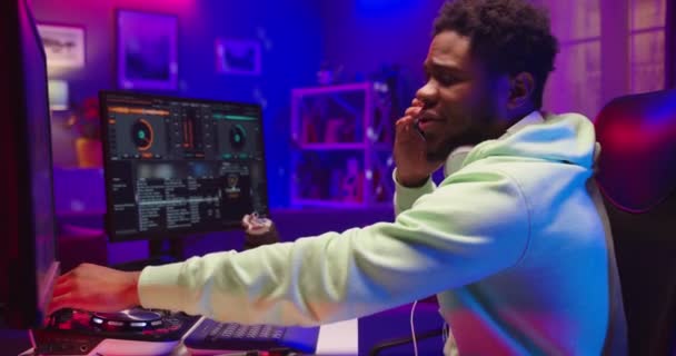 Side view of handsome young African American man dj sitting at desk in home studio in neon light speaking on cellphone while mixing music composing sound on mixer console looking at computer monitor — Stock Video