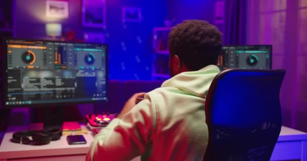 Back view of young African American guy sound engineer in house in neon light sitting at table mixing song hit on mixer controller looking at computer screen, streaming music podcast, dj job, close up — Stock Video