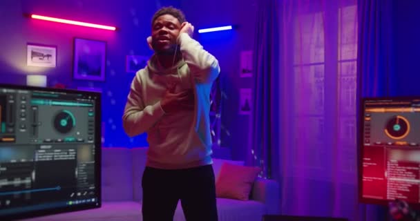 Vreugdevolle Afro-Amerikaanse jonge knappe man staan in donker huis in neon lichten in huis opname studio dansen en ritmisch ontroerende genieten van dans luisteren naar clubmuziek in de woonkamer, dj concept — Stockvideo