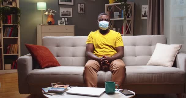 Een zieke volwassen man met een beschermend medisch masker die thuis blijft tijdens een pandemie van het coronavirus. Afro-Amerikaan zit op de bank en kijkt naar de camera in de woonkamer. Isolatie, quarantaineconcept. — Stockvideo