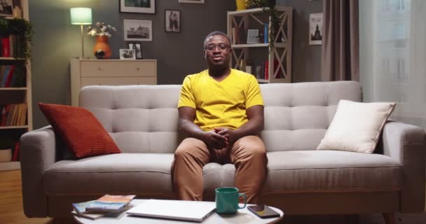 Joven agradable y relajante sentado en el sofá en casa por la noche y mirando a la cámara. Guapo afroamericano en gafas descansando en el sofá y sonriendo en la sala de estar. Concepto de descanso y buen tiempo. — Vídeos de Stock