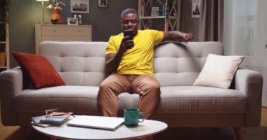Sakin bir genç adam kanepede oturuyor, arkasına yaslanıyor ve evde akıllı telefon kullanıyor. Afro-Amerikalı adam internette geziniyor, video izliyor ve arkadaşlarına mesaj atıyor. Sosyal medya, teknoloji kavramı.