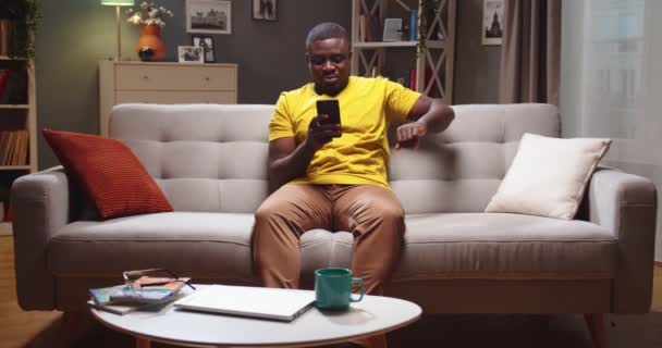 Όμορφος Αφρο-Αμερικανός άντρας χαλαρώνει και ξεκουράζεται στον καναπέ στο σαλόνι. Ενηλίκων τύπος χρησιμοποιώντας smartphone στο μήνυμα, να παρακολουθήσετε βίντεο το βράδυ. Online αγορές, Διαδίκτυο, έννοια του ελεύθερου χρόνου. — Αρχείο Βίντεο
