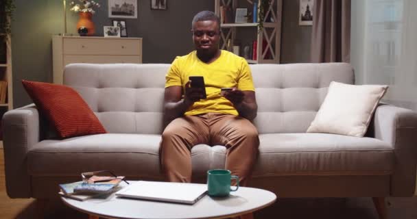 Atrakcyjny mężczyzna siedzący na kanapie i wchodzący numer karty na smartfonie w salonie. Afroamerykanin robi zakupy online, kupuje ubrania i robi zakupy wieczorem. Koncepcja przedsiębiorstwa. — Wideo stockowe