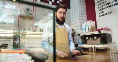 Kahve dükkanında dijital tablet kullanan önlüklü genç bir adamın portresi. Kafkasyalı erkek barista bir aletin üzerine yazıyor, kameraya bakıyor ve gülümsüyor. Küçük işletme, kafe, restoran konsepti.