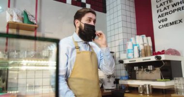 Koruyucu tıbbi maskeli çekici erkek barista telefonda müşteriyle konuşuyor. Yetişkin beyaz bir adam modern şehir kafeteryasında ön sipariş alıyor. İş, covid-19, koronavirüs, karantina konsepti.