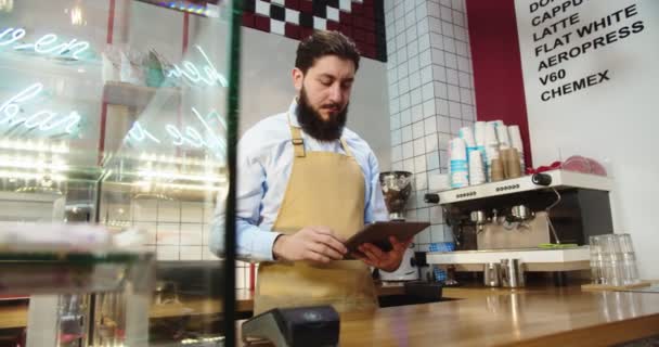 バーの後ろに立って、カフェでタブレットに取り組んでいる良い外観の白人男性。大人の男性コーヒーショップのオーナーは在庫をチェックし、注文を取り、デジタルタブレットで入力します。ビジネス、サービス. — ストック動画