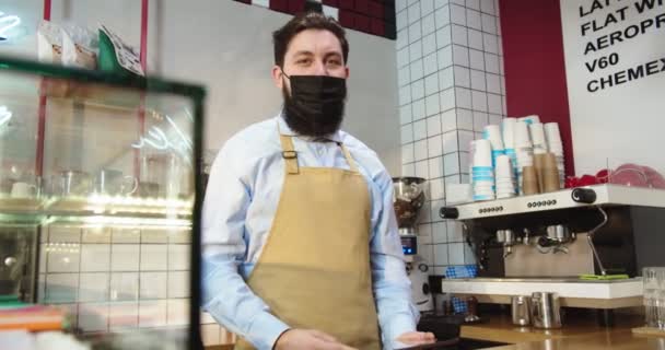 Portret van een charmante man met een beschermend medisch masker achter de bar in het café. Jonge blanke mannelijke barista draagt schort, kijkt naar de camera en glimlacht in de koffieshop. Bedrijfs-, quarantaineconcept. — Stockvideo