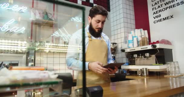 Profesjonalny młody barista pracujący na tablecie, przyjmujący zamówienia i sprawdzający zapasy w kawiarni. Biały właściciel kawiarni pisze na gadżecie, patrzy w kamerę i uśmiecha się. Koncepcja przedsiębiorstwa. — Wideo stockowe