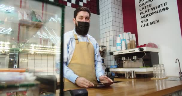 Profesionální mužský barista v ochranné masce pracující v městské kavárně. Dospělý muž používá digitální tablet, dělá si poznámky, dívá se do kamery a usmívá se. Covis-19, pandemie, koncepce karantény. — Stock video