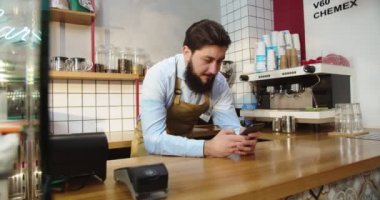 Profesyonel erkek barista barın arkasında duruyor ve kafede akıllı telefon kullanıyor. Genç beyaz bir adam arkadaşlarıyla sohbet ediyor ve sosyal medyaya bakıyor. Kahve dükkanı, iş yeri, teknoloji..