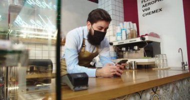 Tıbbi koruyucu maskeli yakışıklı küçük işletme sahibinin portresi. Genç beyaz adam emirler almak için akıllı telefonu kullanıyor, daktilo ediyor ve sosyal medyayı inceliyor. Teknoloji konsepti.