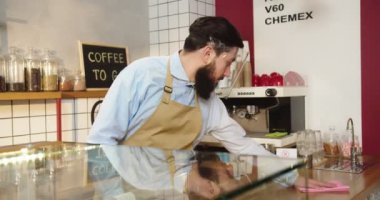 Yüz kalkanı takmış yakışıklı bir adamın portresi kafedeki bar yüzeyini dezenfekte ediyor. Yetişkin erkek barista masayı siliyor, kameraya bakıyor ve gülümsüyor. Sosyal mesafe tabelasını koru. Virüs, karantina.