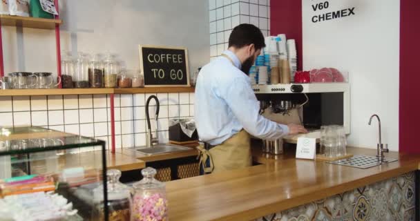 Hårt arbetande ung kille som jobbar och torkar av kaffemaskinen på caféet. Vit manlig barista desinficerar, desinficerar arbetsplatsen, tittar på kameran och ler. Rena kassaskåpet. Viruskoncept. — Stockvideo