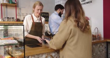 Kahve parasını ödeyen güzel bayan müşteri NFC teknolojisini telefonla kullanıyor ve sipariş alıyor. Kadın kafe çalışanı terminalden bayana uzatıyor. Temassız ödeme, teknoloji konsepti.