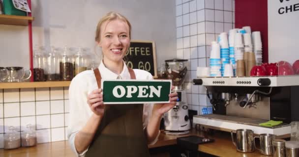 バーの後ろに立って新しい喫茶店でOPENの看板を見せる楽しい女。小さなカフェビジネスを開始成功した若いバリスタ。起業家精神、キャリア、人生の目標コンセプト. — ストック動画