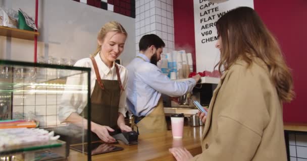 Alımlı genç bayan barista müşteriye ulaşılamayan ödeme terminalini tutuyor. Çekici beyaz kadın telefonla ödeme yapıyor ve kahve fincanı siparişi alıyor. İş, NFC teknoloji kavramı. — Stok video