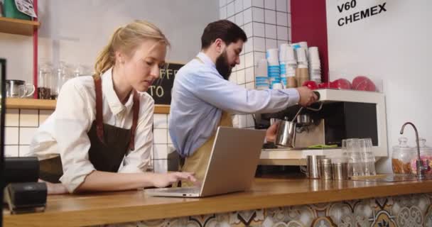 Ενηλίκων γυναίκα ιδιοκτήτης επιχείρησης καφέ λαμβάνοντας παραγγελίες και τον έλεγχο των αποθεμάτων στο φορητό υπολογιστή. Λευκός μπάρμαν εργάζεται και φτιάχνει καφέ στο παρασκήνιο. Entreprenuership, έννοια τεχνολογιών. — Αρχείο Βίντεο