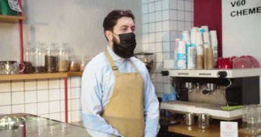 Önlük giyen ve tezgahın arkasında duran çekici genç adam. Koruyucu tıbbi maskeli beyaz erkek kahve dükkanı sahibi kameraya bakıyor. Coronavirus, pandemik konsept.
