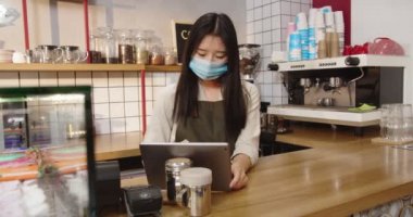 Tablet ve daktilo kullanarak koruyucu maske takan profesyonel bayan barista. Genç Asyalı kadın, kahve dükkanındaki aletlerle internetten müşteri siparişi alıyor. Coronavirus, pandemik konsept.