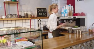 Modern kahvehanede çalışan güzel bir bayan barista kahve makinesinde içecek yapıyor. Kafkasyalı genç bir kadın içki bardağını alıyor, barın üstüne çıkıp kameraya bakıyor ve gülümsüyor. İş dünyası.