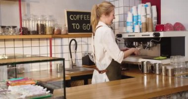 Kafe barında çalışan güzel bir bayan barmen, içki hazırlıyor. Kafkasyalı genç bir kadın kahve makinesi kullanıyor, içecek alıyor, masaya oturuyor, gülümsüyor ve kameraya bakıyor. Hizmet, iş.