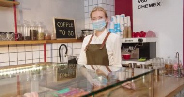 Barın arkasında duran tıbbi koruyucu maskeli güzel bayan barmen. Önlük giyen genç beyaz kadın kameraya bakıyor ve gülümsüyor. İş, karantina, virüs konsepti.