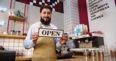 Şehir kafeteryasında çalışan ve barın arkasında duran beyaz bir adam. Açık tabela tutan genç erkek barista kameraya bakıyor ve kafede gülümsüyor. İş, başlangıç, başarı kavramı.