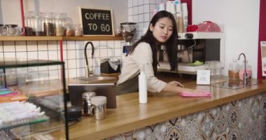 Önlüklü çekici bayan barmen kafede alkolle bardan çıkıyor. Yetişkin Asyalı kadın masa yüzeyini sterilize ediyor, kameraya bakıyor ve kafede gülümsüyor. Temiz kasa. Virüs konsepti.