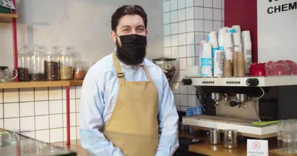 Πορτρέτο ενός όμορφου Καυκάσου που δουλεύει σε σύγχρονη καφετέρια. Νεαρό αρσενικό barista σε ιατρική προστατευτική μάσκα κοιτάζοντας κάμερα και χαμογελώντας στο καφέ. Επιχειρήσεις, υπηρεσίες, concept covid-19. — Αρχείο Βίντεο