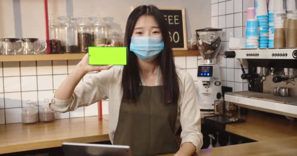バーの後ろに立ってコーヒーショップでクロマキーを示す美しいアジアの女性。緑の画面でスマートフォンを見て医療用マスクの若い女性バーテンダー。水平位置. — ストック動画