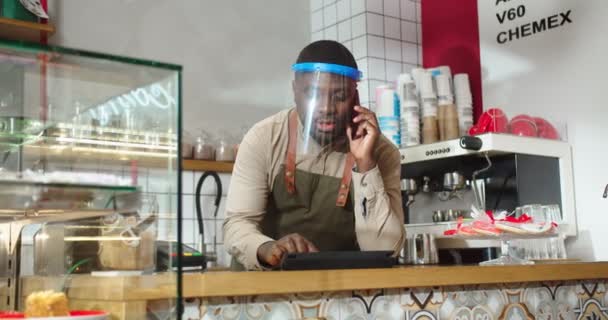 Çalışkan erkek barista müşterinin siparişini alıyor ve dizüstü bilgisayara yazıyor. Yüz koruyucu vizörlü Afro-Amerikalı bir adam müşteriyle telefonda konuşuyor ve cihaz üzerinde yazıyor. Kahve dükkanı, iş yeri.. — Stok video