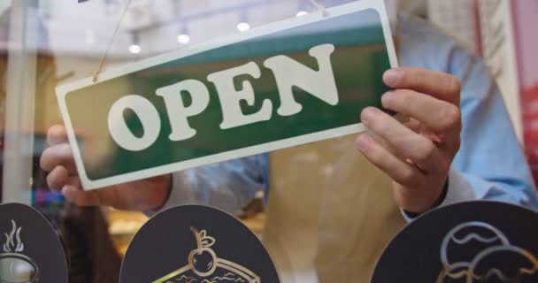 Vooraanzicht van koffieshop deur tijdens pandemie periode. Jonge blanke mannelijke barista in schort sluiten cafe als gevolg van crisis en flipping teken van Gesloten naar OPEN. Covid-19, afsluitmaatregelen.. — Stockvideo