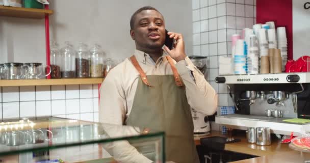 Bájos férfi afro-amerikai barista beszél telefonon a kávézóban. Fiatal fickó kötényes egyenruhában, szoknyaszünetet tart és hívja a barátját. Kommunikáció, technológiák, eszközök, üzleti koncepció. — Stock videók