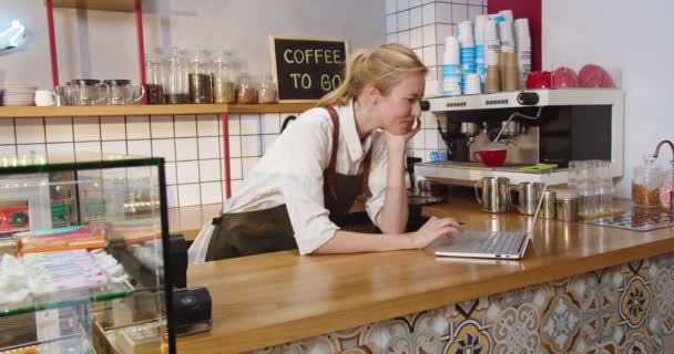 Charmante kaukasische Dame, die am Laptop im städtischen Café arbeitet. Junge Café-Inhaberin tippt, prüft Lager, nimmt Bestellungen entgegen, blickt in die Kamera und lächelt. Business, Technologien Konzept. — Stockvideo