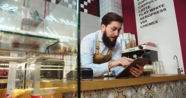 Homem bonito trabalhando, digitando, verificando estoque e tomando pedidos on-line em tablet. Homem adulto caucasiano barista inclinado no bar e usando gadget digital no café. Negócio, conceito de profissão. — Vídeo de Stock