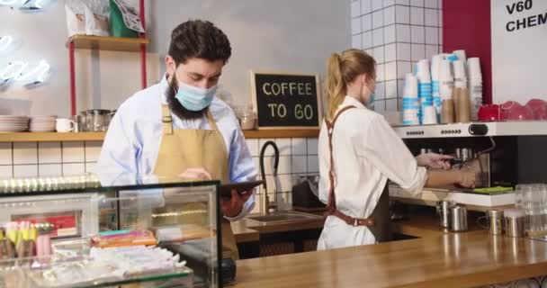 Pohledný běloch přijímá objednávky na stůl a krásná mladá žena dělá kávu. Příjemný personál kavárny v lékařských maskách, dívá se do kamery a usmívá se. Virus, pandemický koncept. — Stock video